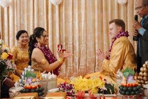 Wedding ceremony scene with floral decorations and fruit displays as participants exchange garlands in a traditional setting.