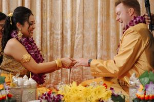 Two people exchange rings during a traditional Hawaiian wedding ceremony with colorful leis and flowers surrounding them.
