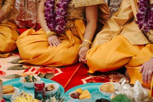 Saffron-robed individuals sitting cross-legged on a red mat with purple leis and traditional offerings at a ceremony.