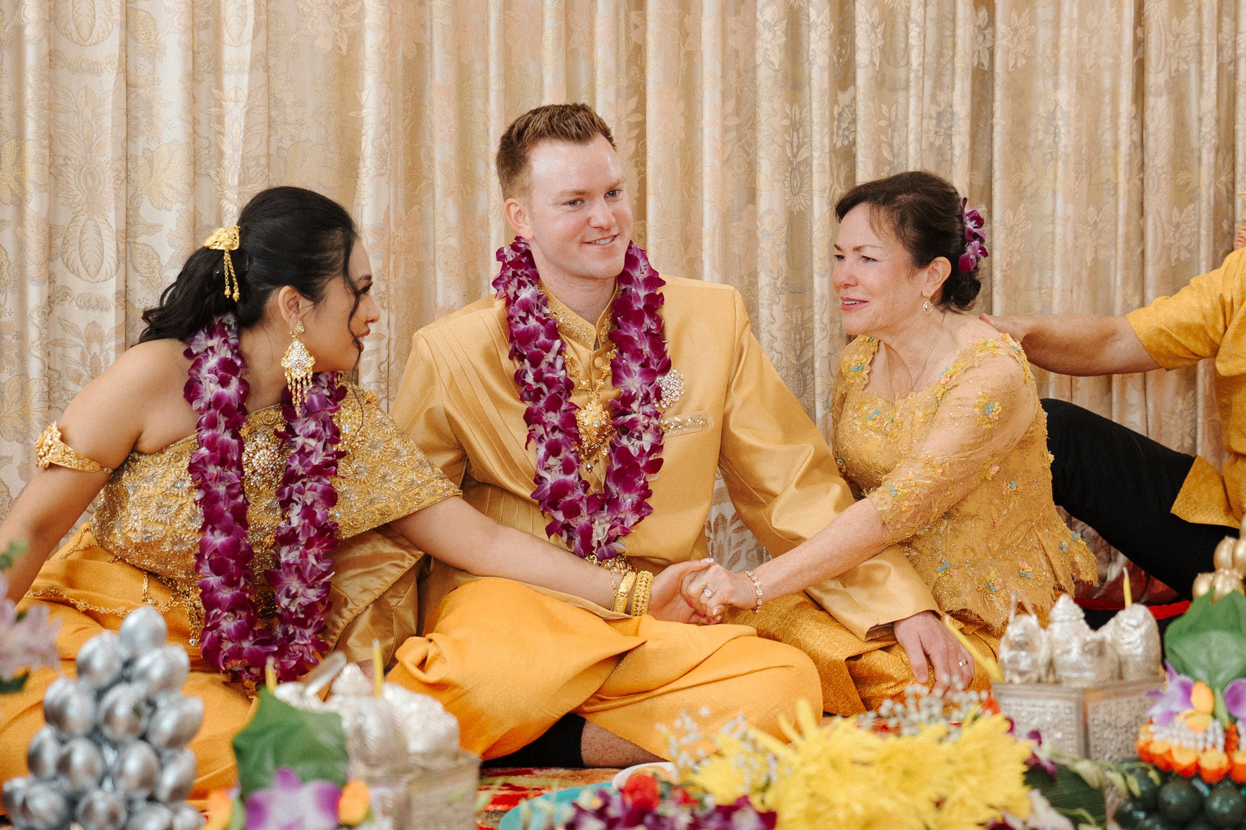 Traditional ceremony participants in yellow and orange attire seated at a long table with purple floral decorations.