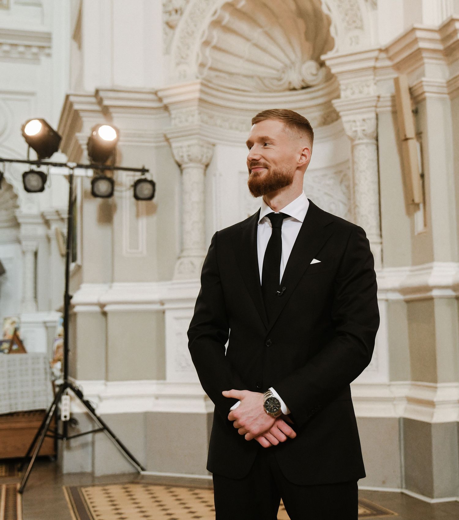 Person in black suit standing in elegant venue with ornate white architectural details and vintage lighting.