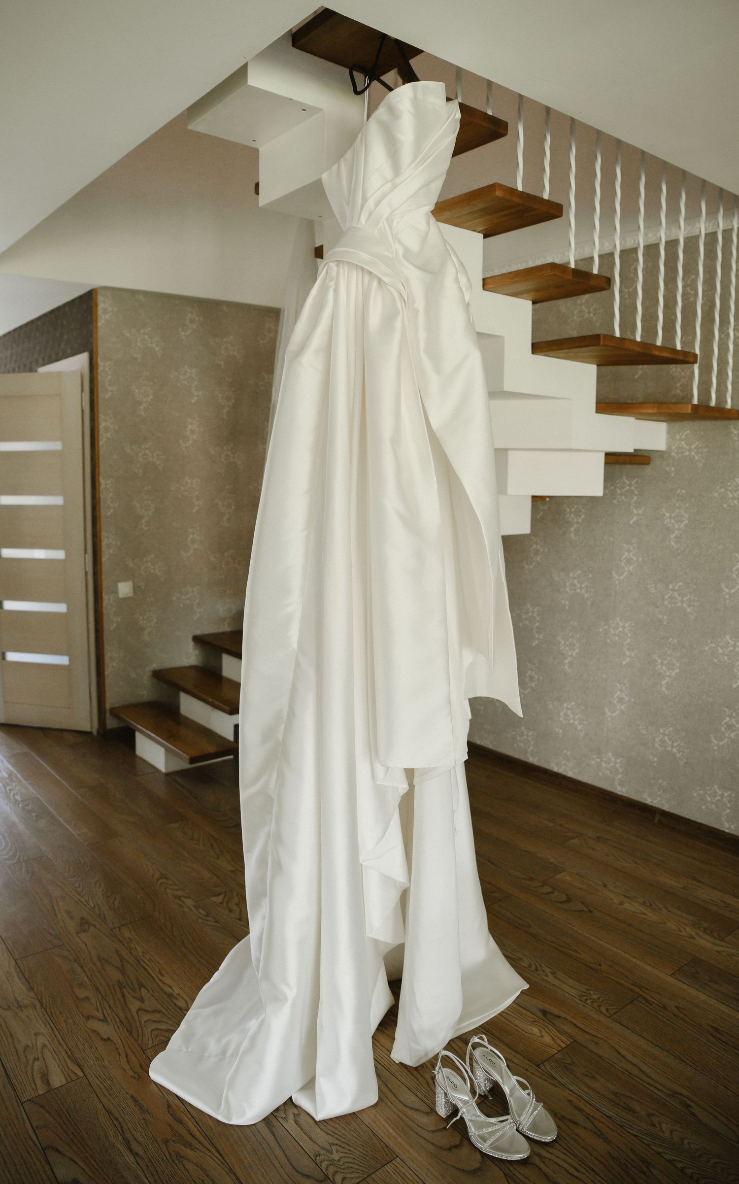 An elegant white wedding dress hangs on display with matching shoes underneath by a modern wooden staircase.