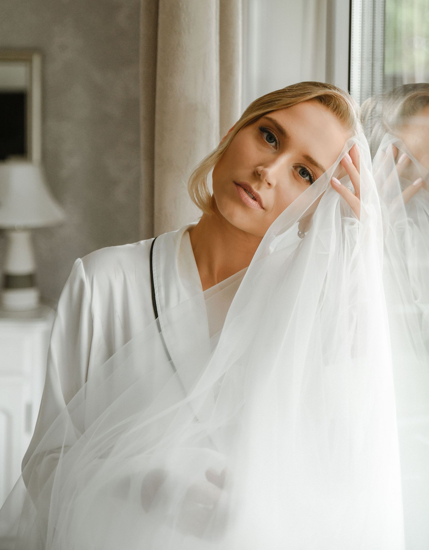 Figure in white robe poses behind sheer curtains in dreamy indoor setting.