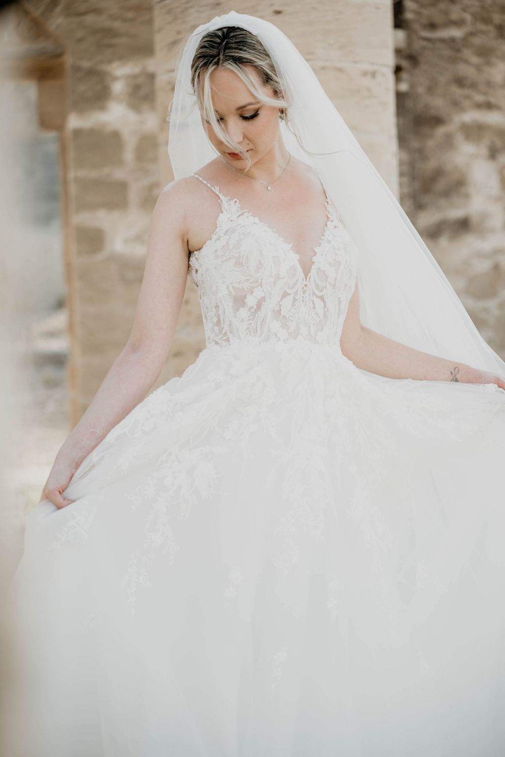 Elegant white wedding gown with lace bodice and flowing tulle skirt paired with delicate veil against stone wall backdrop.