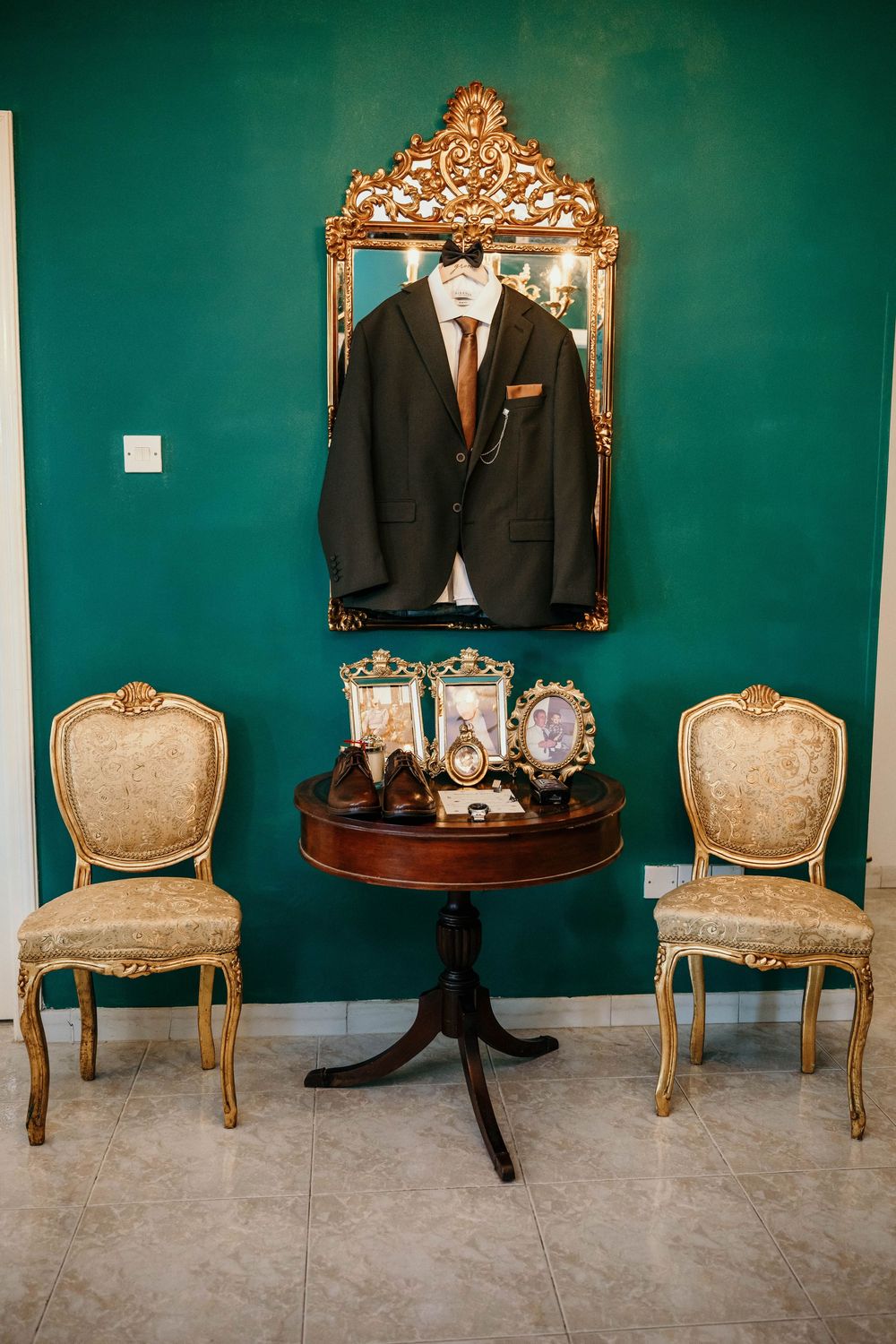 Elegant vintage setup with two beige chairs, ornate gold mirror and dark suit on emerald green wall with round table display.