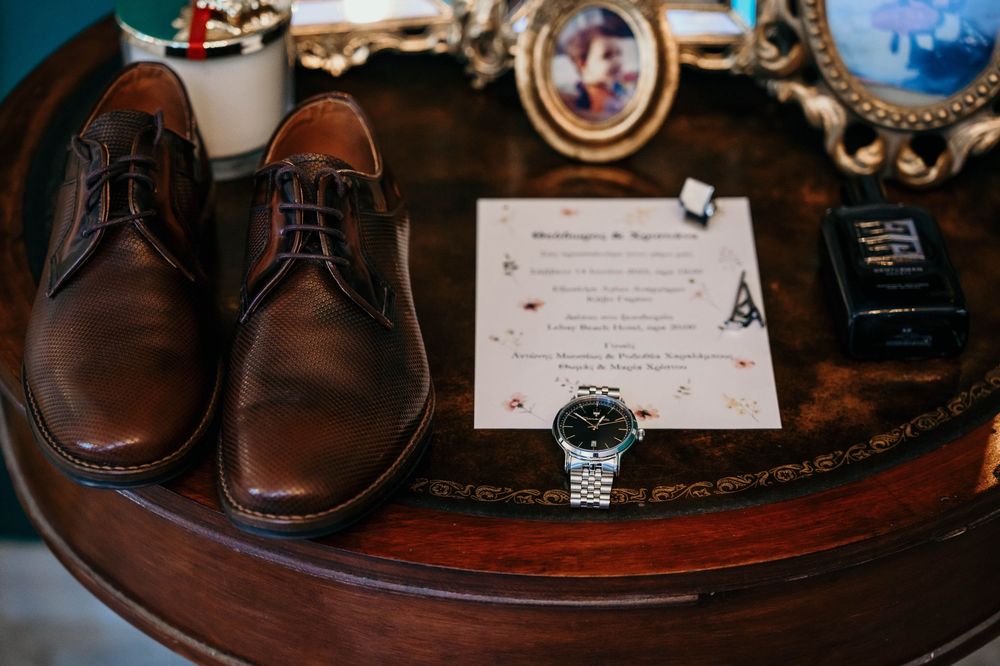 Brown leather dress shoes, watch, and wedding invitation displayed on vintage wooden table with ornate decor.