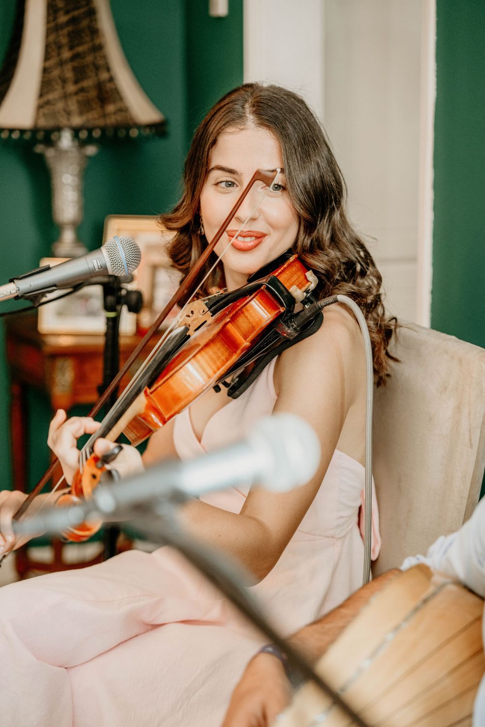 A musician performs with a violin while seated in a room with green walls and a decorative lamp.