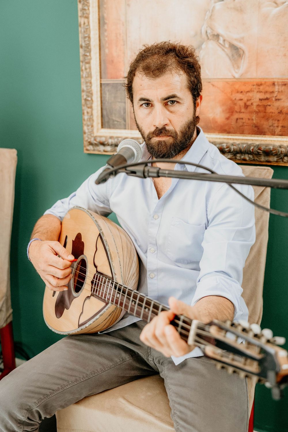 A musician plays a traditional stringed instrument while seated in front of a teal wall with artwork.