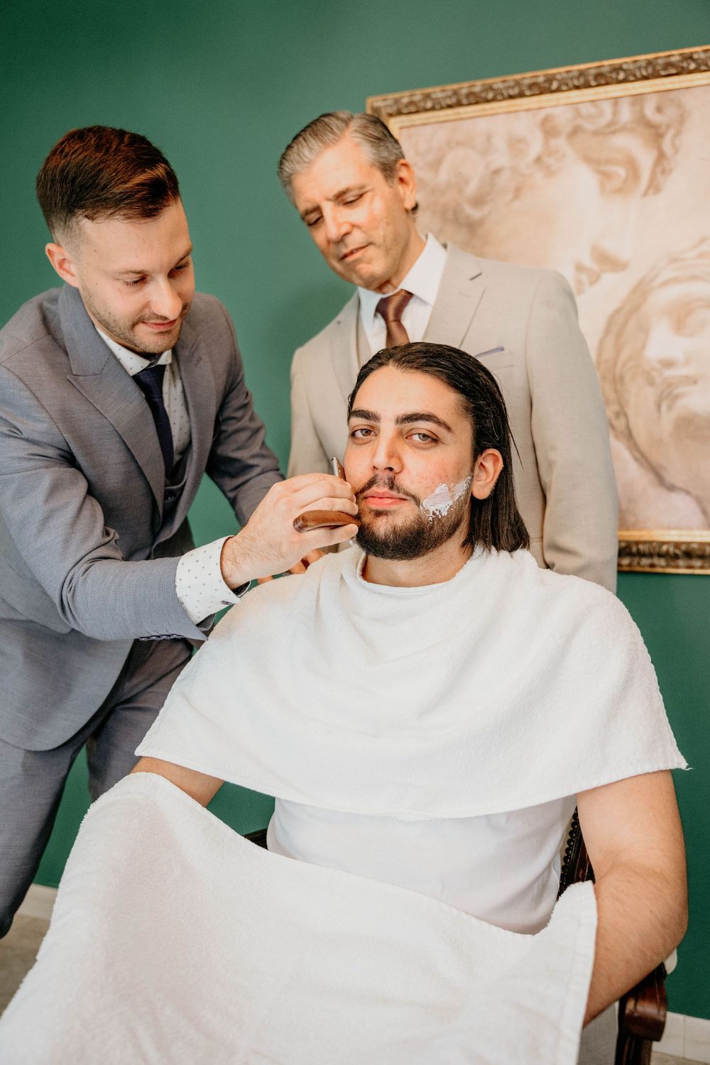 Two professional barbers work together to groom a client wearing a white cape in an elegant salon setting.