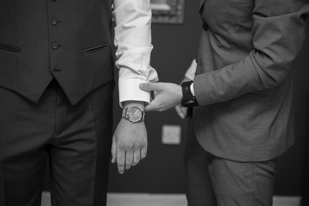 A black and white closeup of two people wearing suits and watches holding hands together.