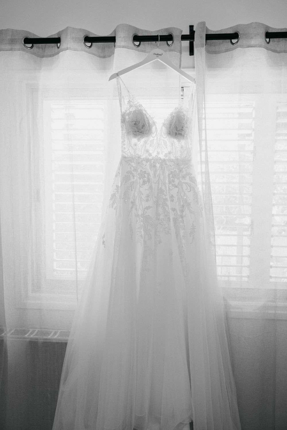 Three ethereal white wedding dresses hang against a light-filled window with sheer curtains creating a dreamy silhouette.