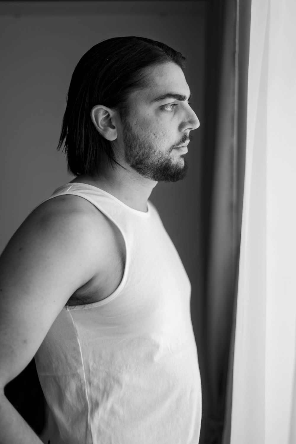 Black and white profile portrait of someone with dark hair and beard wearing a white tank top by a window.