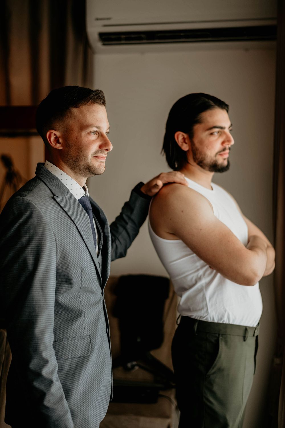 Two people stand side by side indoors, one wearing a grey suit and one in a white tank top and green pants.