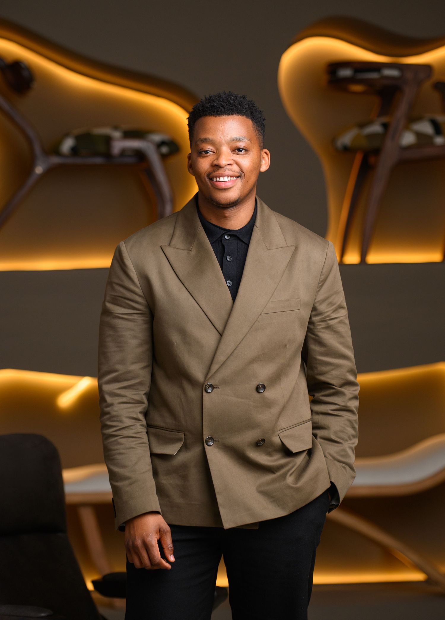 Professional portrait showing a person in a khaki double-breasted blazer and black shirt against a curved wall with warm lighting.