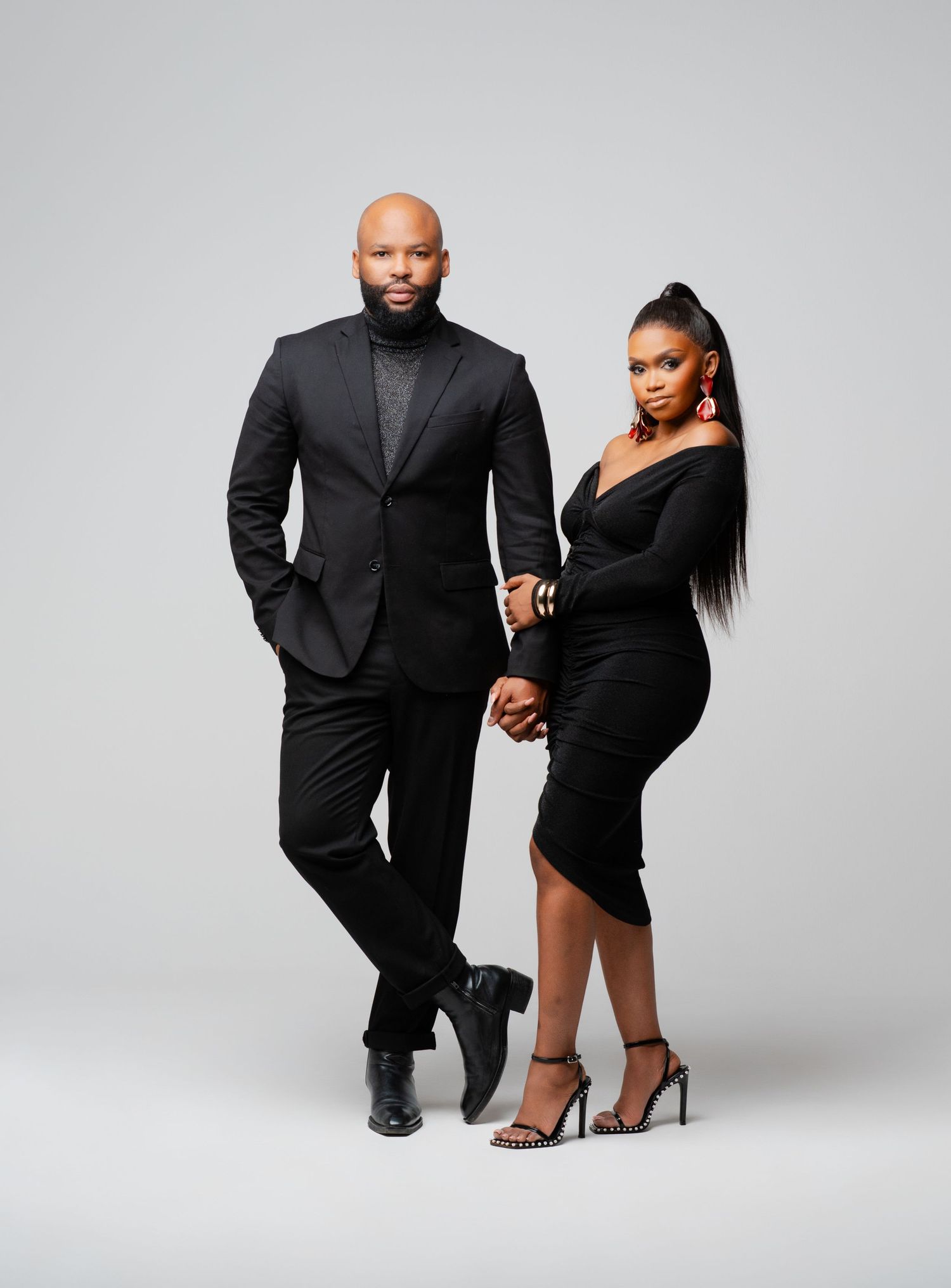 A stylish couple poses together in black formal attire against a light gray studio backdrop.