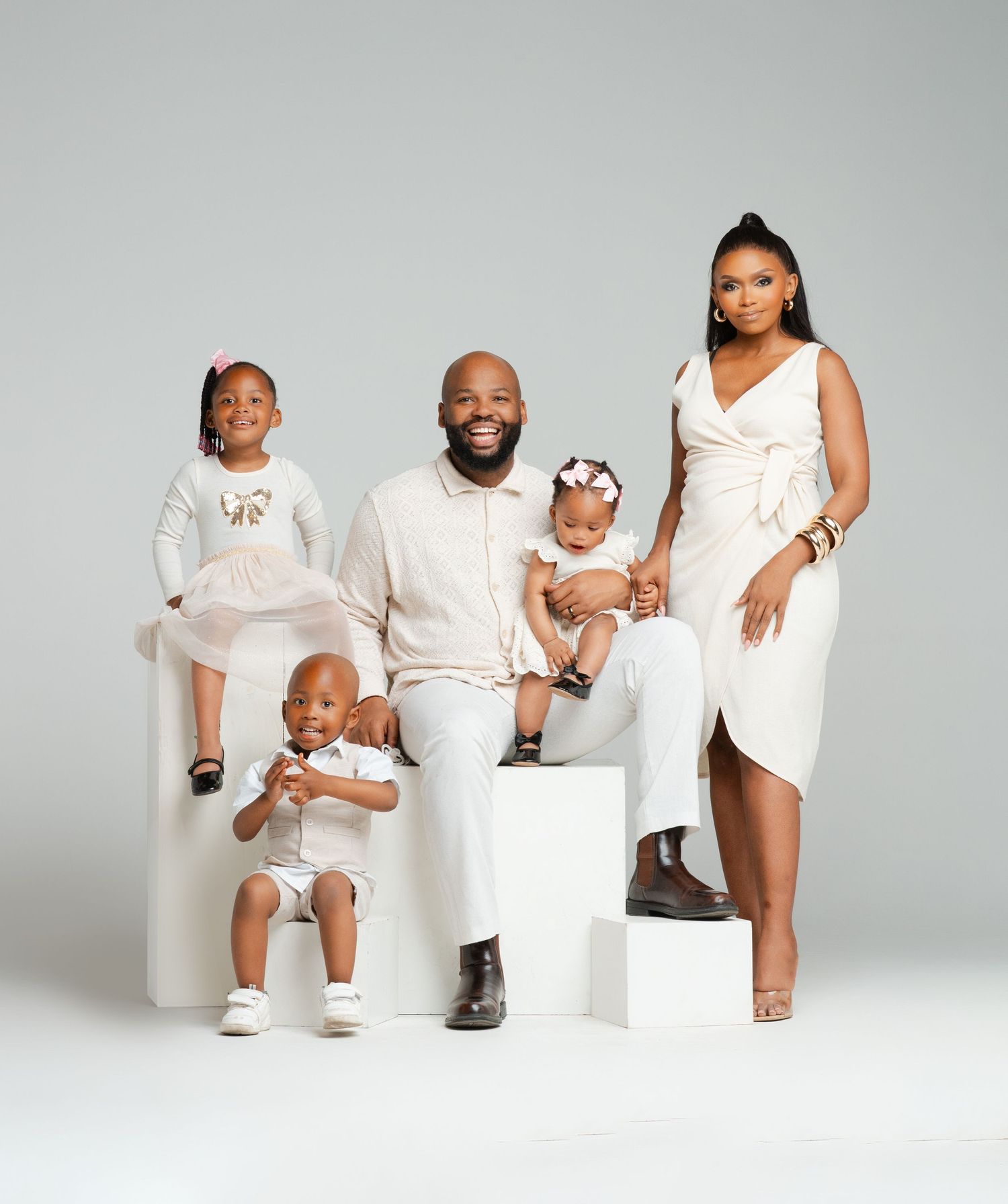 A family of five wearing coordinated white outfits poses together on white steps against a gray backdrop.
