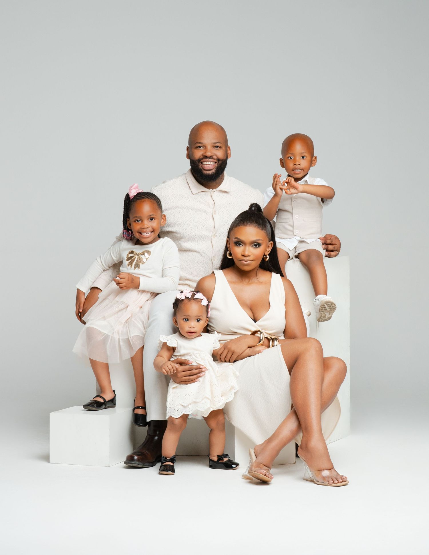 A family dressed in matching white outfits sits together on white platforms for a studio portrait.