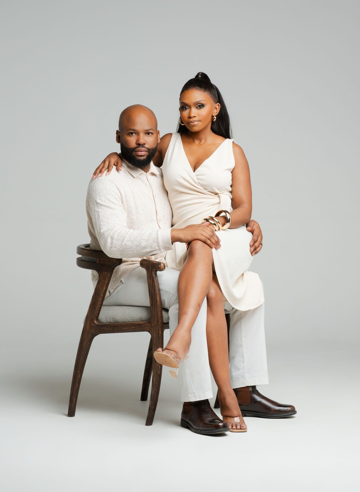 A couple in elegant white outfits poses intimately on wooden chairs in a minimalist studio setting.