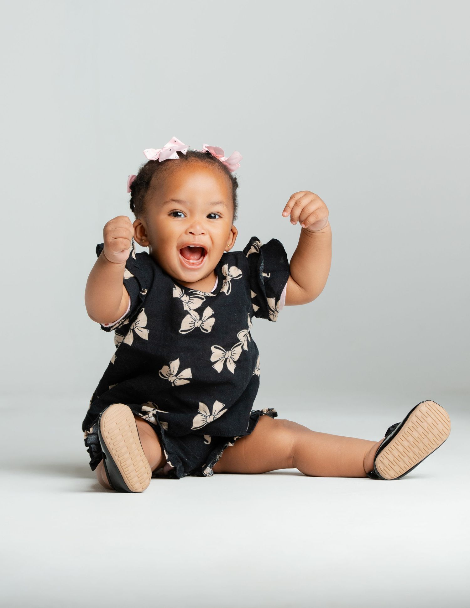 A baby in a black dress with white butterfly prints sits and plays on a white studio backdrop.