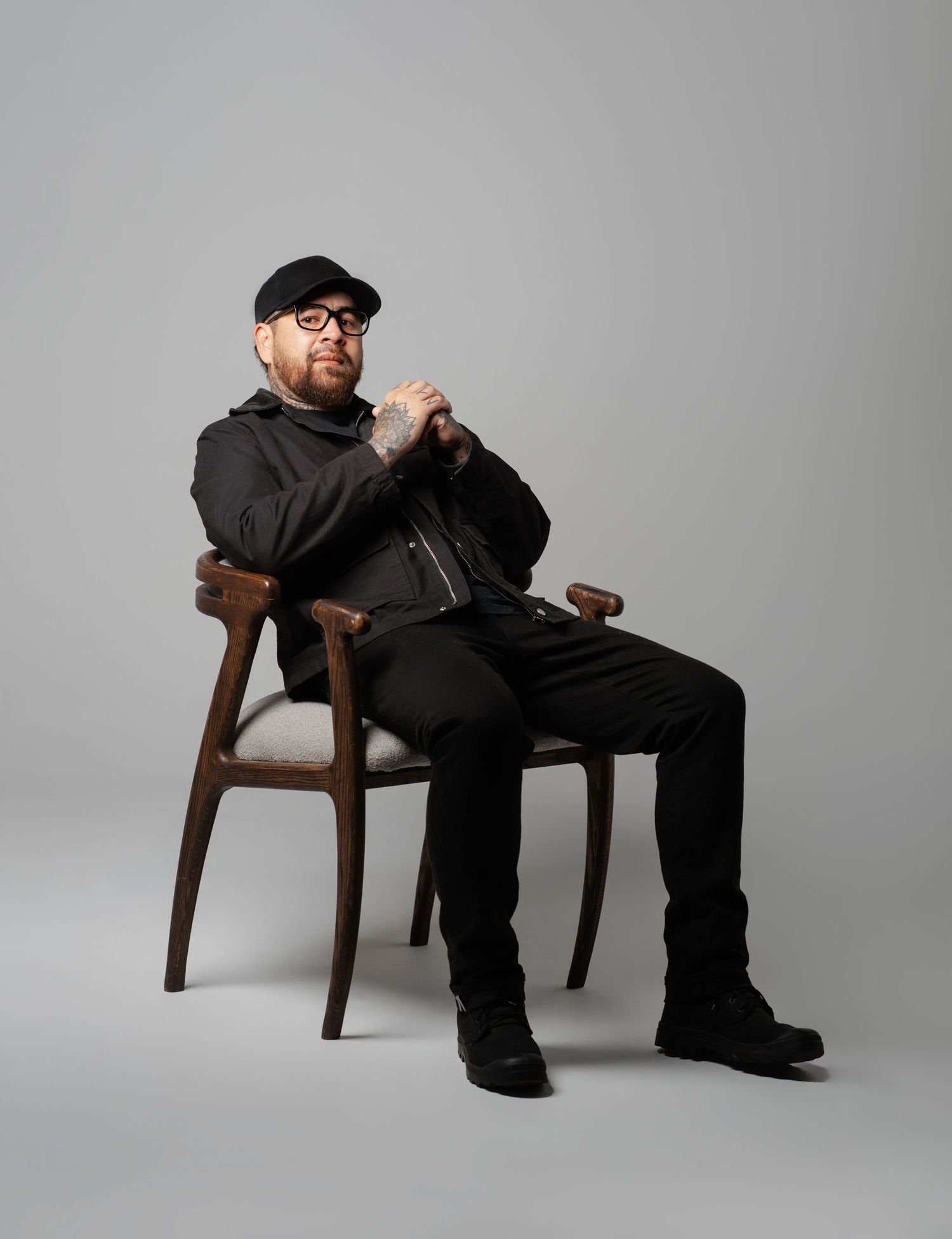 A figure wearing black clothing and glasses sits in varying casual poses on a wooden chair against a gray backdrop.