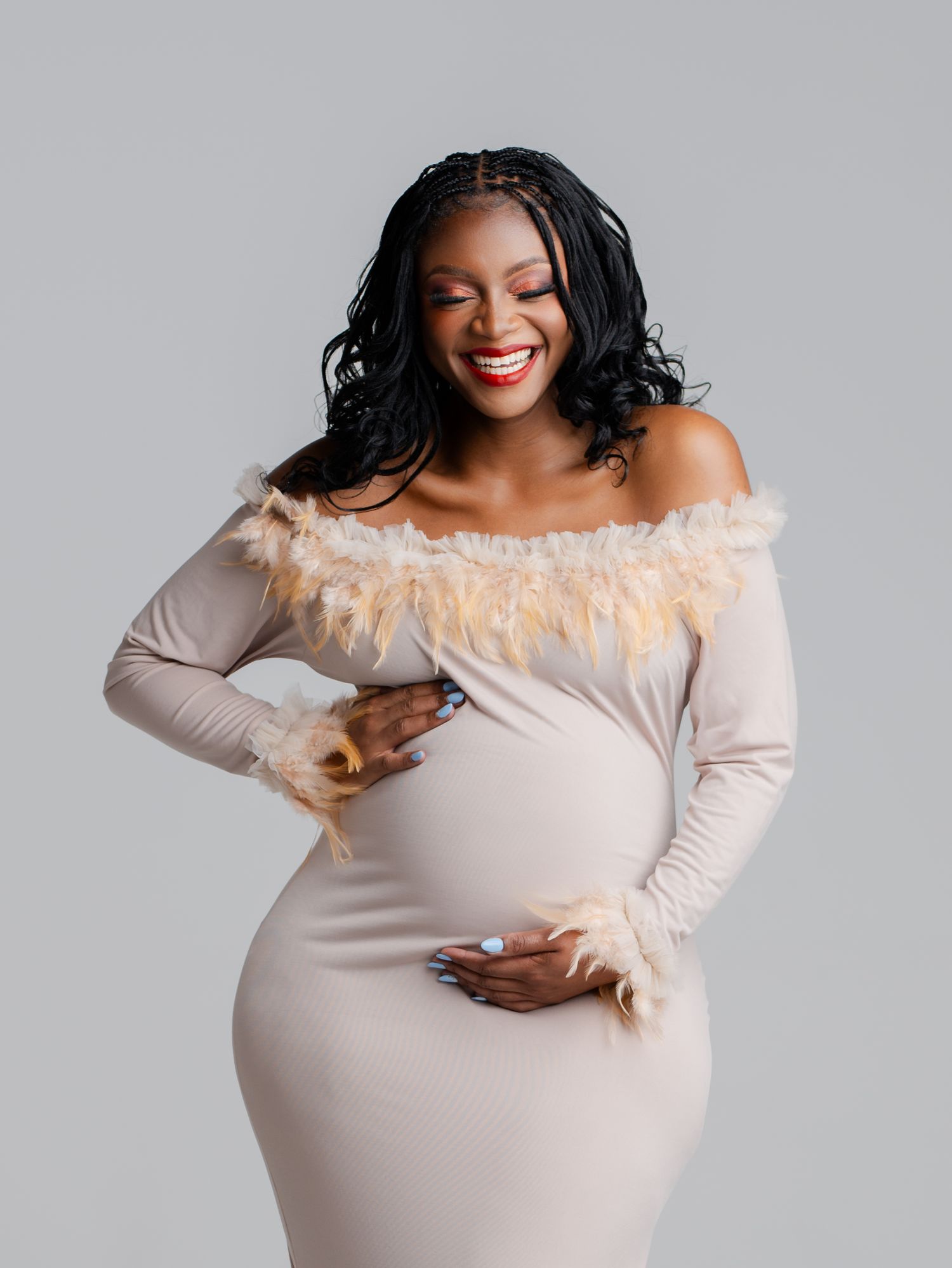 Elegant maternity photo series showing expectant mother in cream off-shoulder dress with feather trim against gray backdrop.