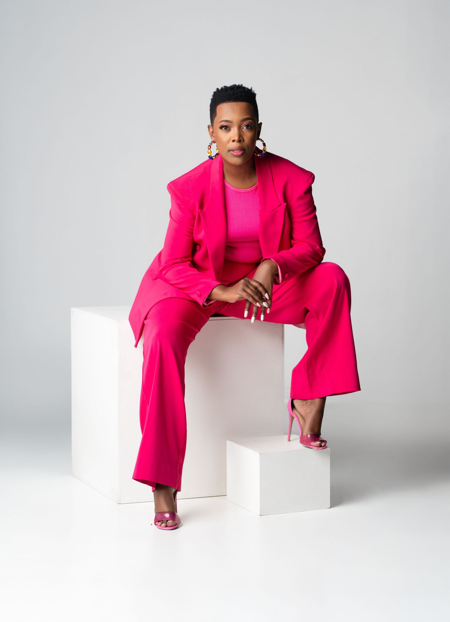 A person wearing a vibrant hot pink pantsuit and matching top poses confidently on white geometric blocks against a gray backdrop.