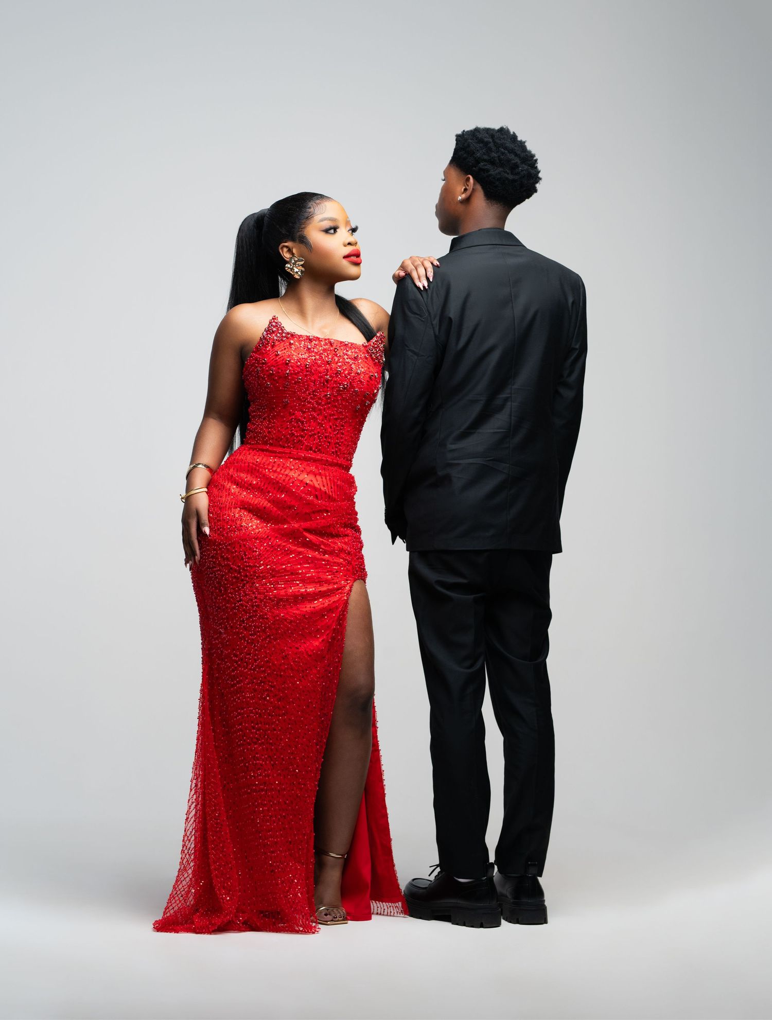 A couple poses together in a red sequined gown and black suit during a formal photo shoot against a light background.