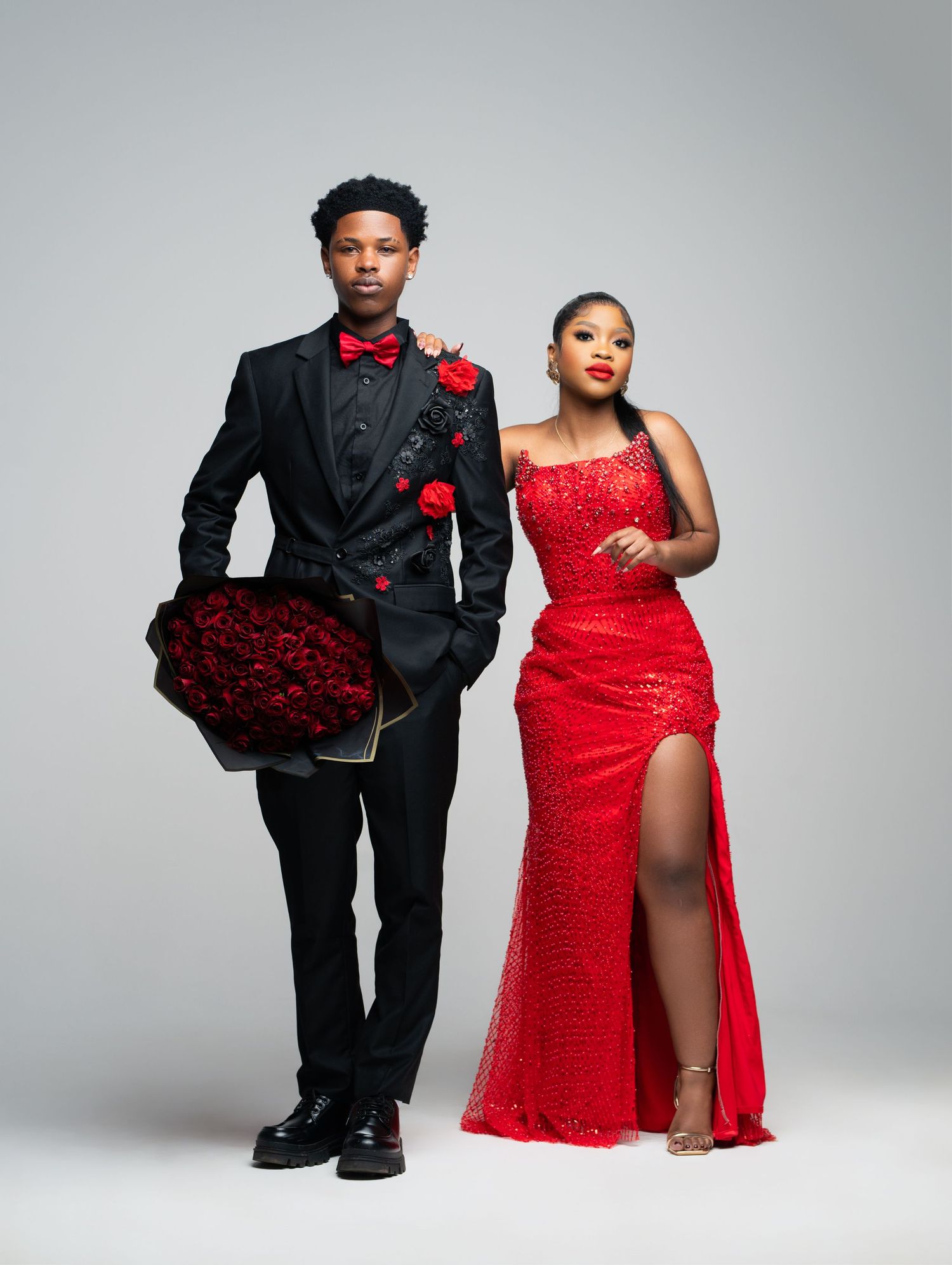 A couple poses in elegant black and red formal attire for a romantic photoshoot against a light gray background.