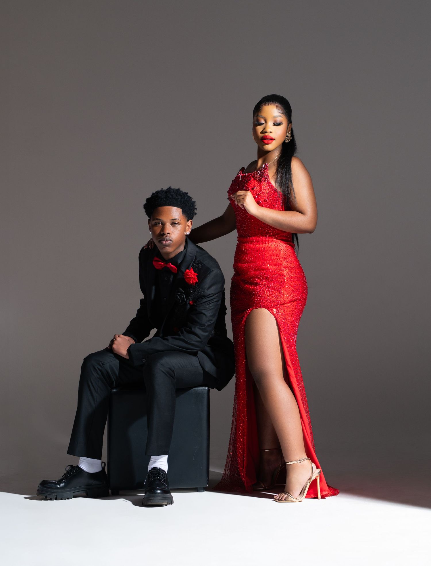 A couple in formal attire poses for a prom photo, with him in a black suit and her in a stunning red lace gown.
