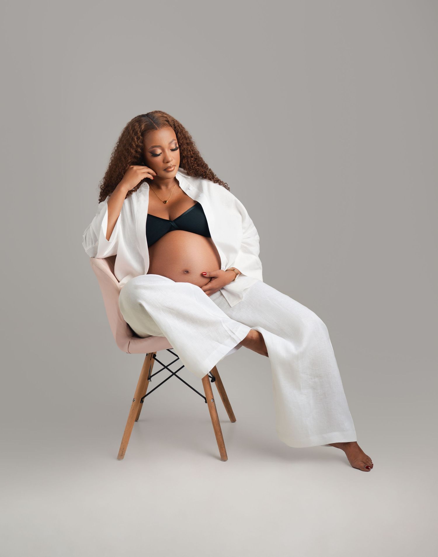 A person in a white robe and black bra sits relaxed on a modern chair against a gray studio backdrop.