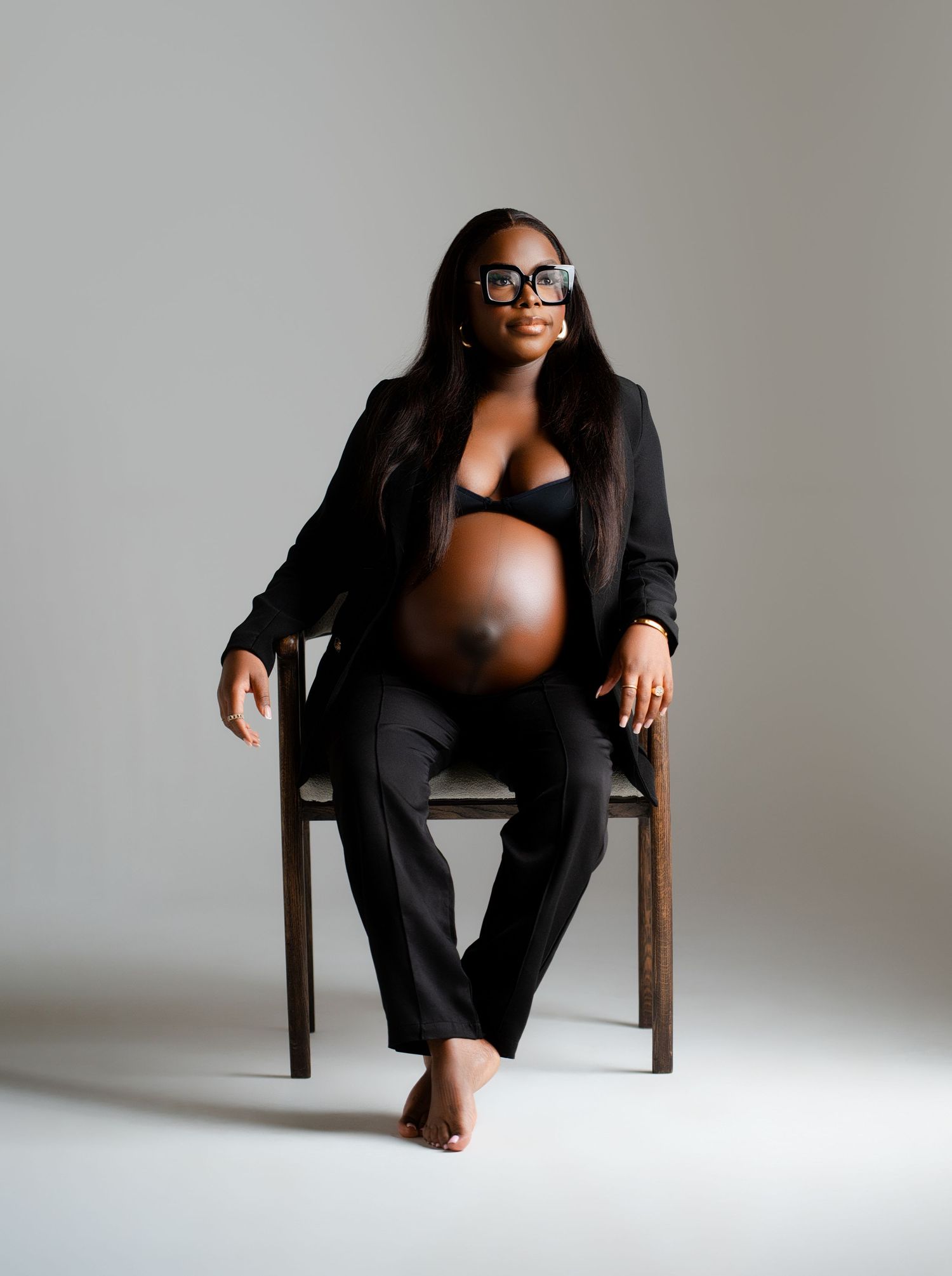 Stylish maternity portrait featuring a black blazer and pants against a minimalist gray backdrop.