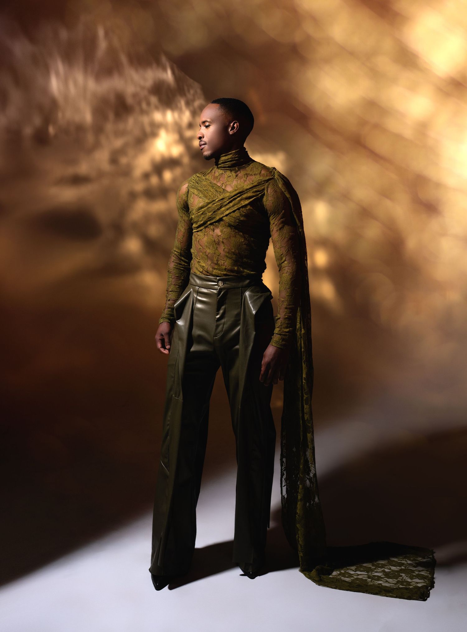 A figure stands dramatically in an olive green ensemble with flowing cape against a moody golden-brown backdrop.