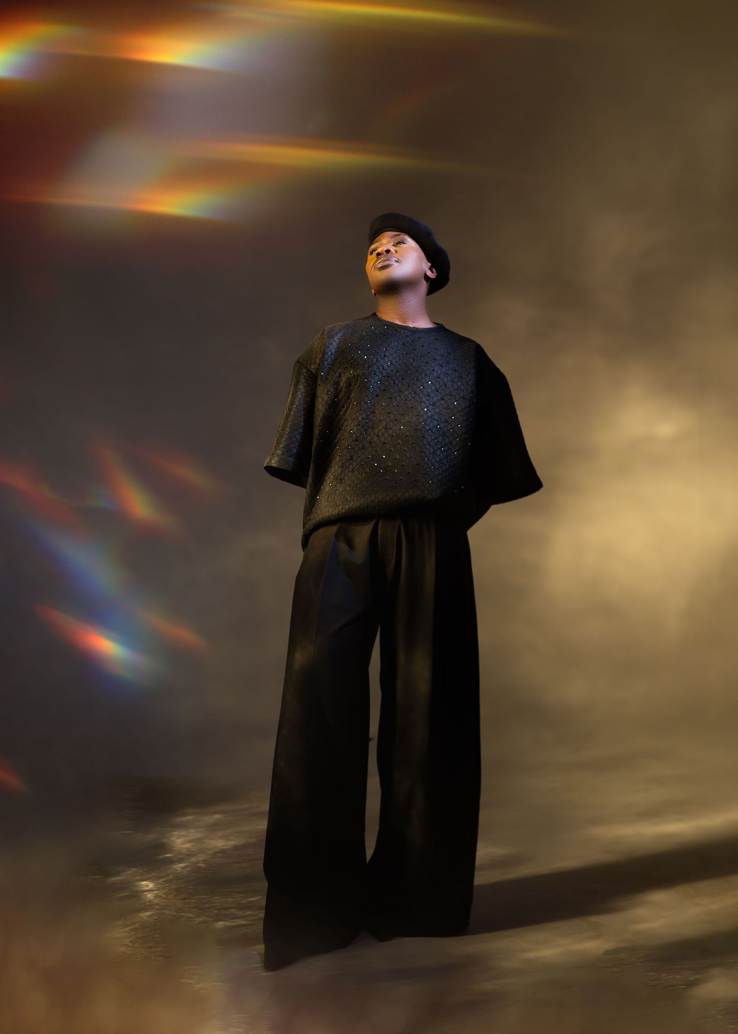 A figure in a dark flowing garment stands silhouetted against a dramatic sky with lens flare effects and rainbow colors.