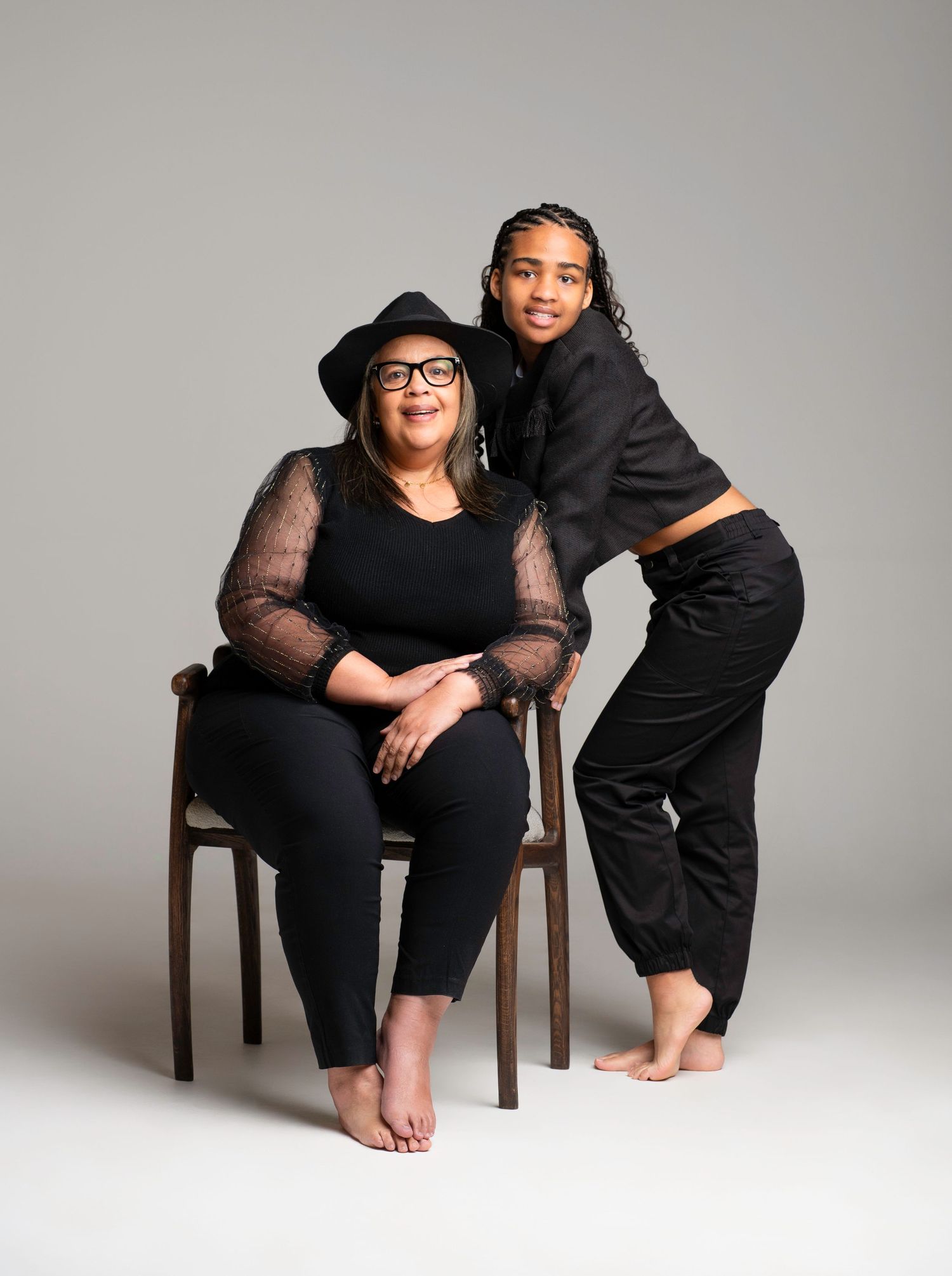 Four minimalist portrait photos show a person in black clothing seated on a wooden chair against a light gray studio backdrop.