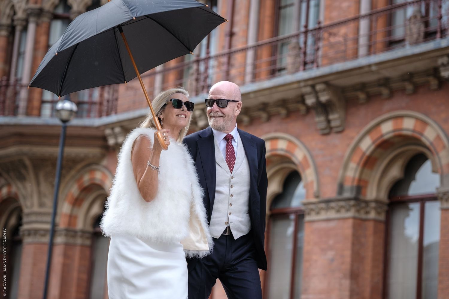 Wedding couple holds black umbrella while posing outside historic brick building in romantic rainy day scene.