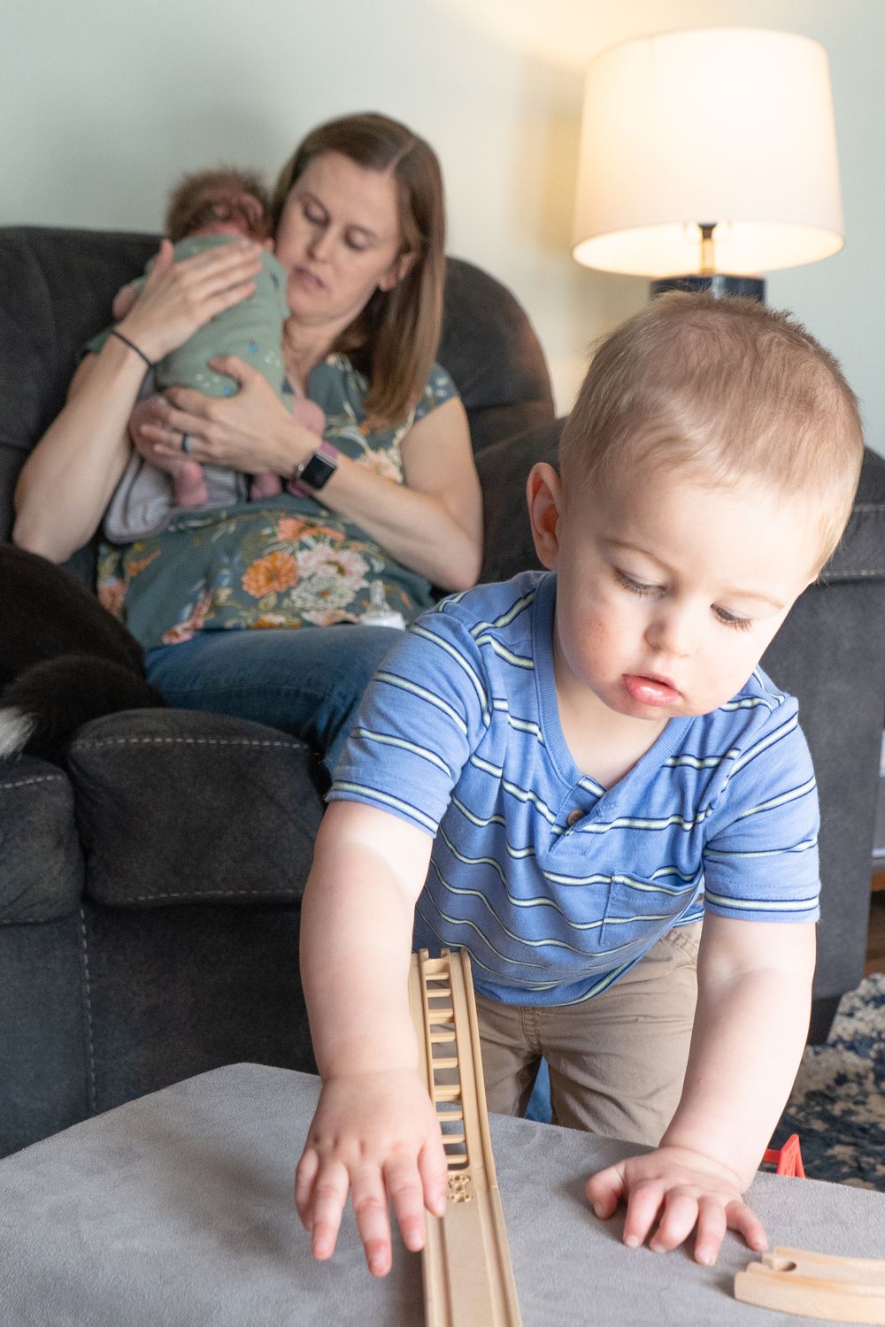 A child plays with wooden blocks on a table while others relax on a dark couch in a cozy living room setting.