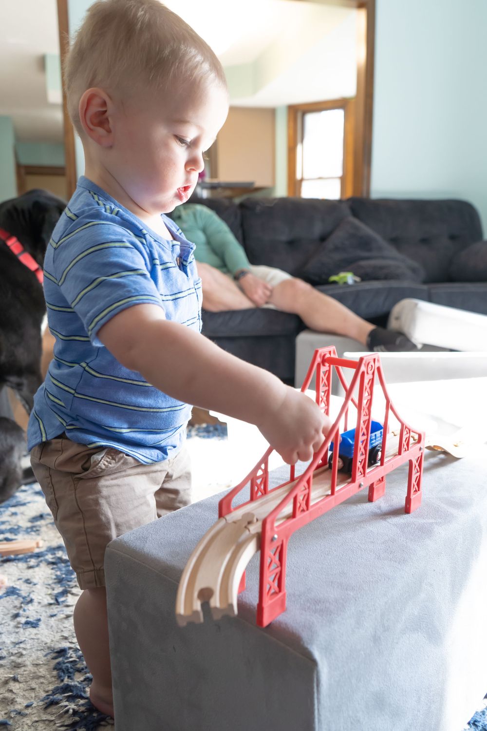 Small child plays with a red model bridge toy sitting on a gray couch cushion.