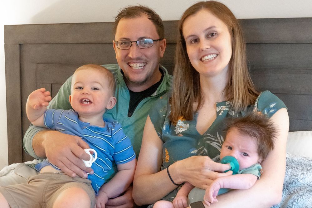 Family portrait in a casual home setting with two adults wearing glasses and casual clothing sitting with two young children.