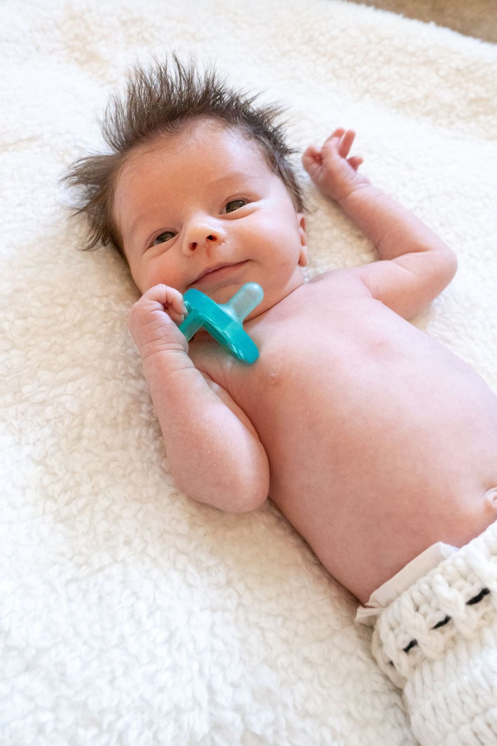 Small baby lying on soft white blanket holding a green pacifier while stretching arms out.