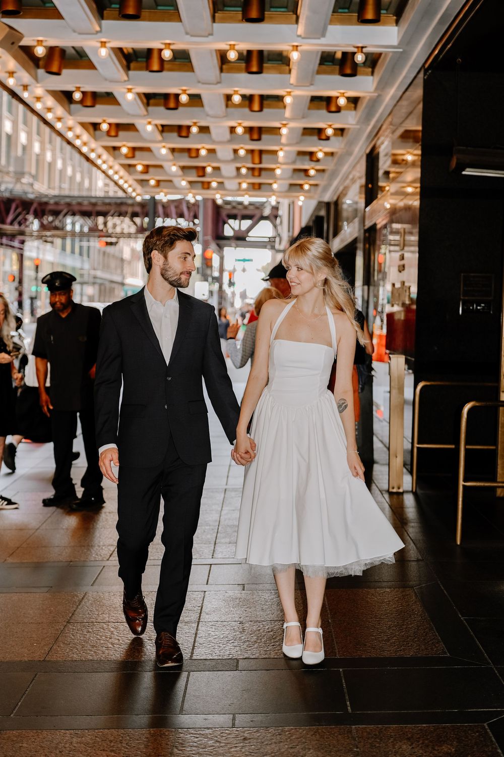 A couple walks together through a brightly lit indoor corridor holding hands, with the bride wearing a short white dress.