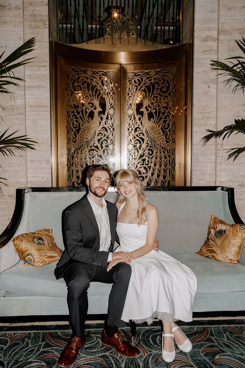A couple sits together on an elegant couch with ornate patterns and palm plants in the background.