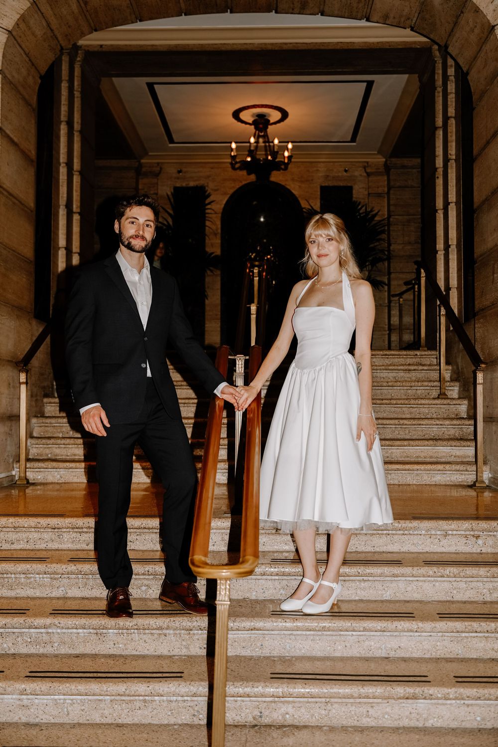 Wedding couple poses elegantly on grand marble staircase in classic black and white attire.