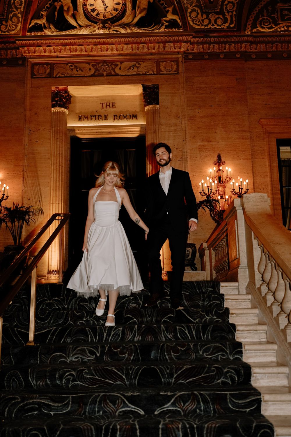 A couple descends an elegant staircase with ornate railings and chandeliers in a historic building.