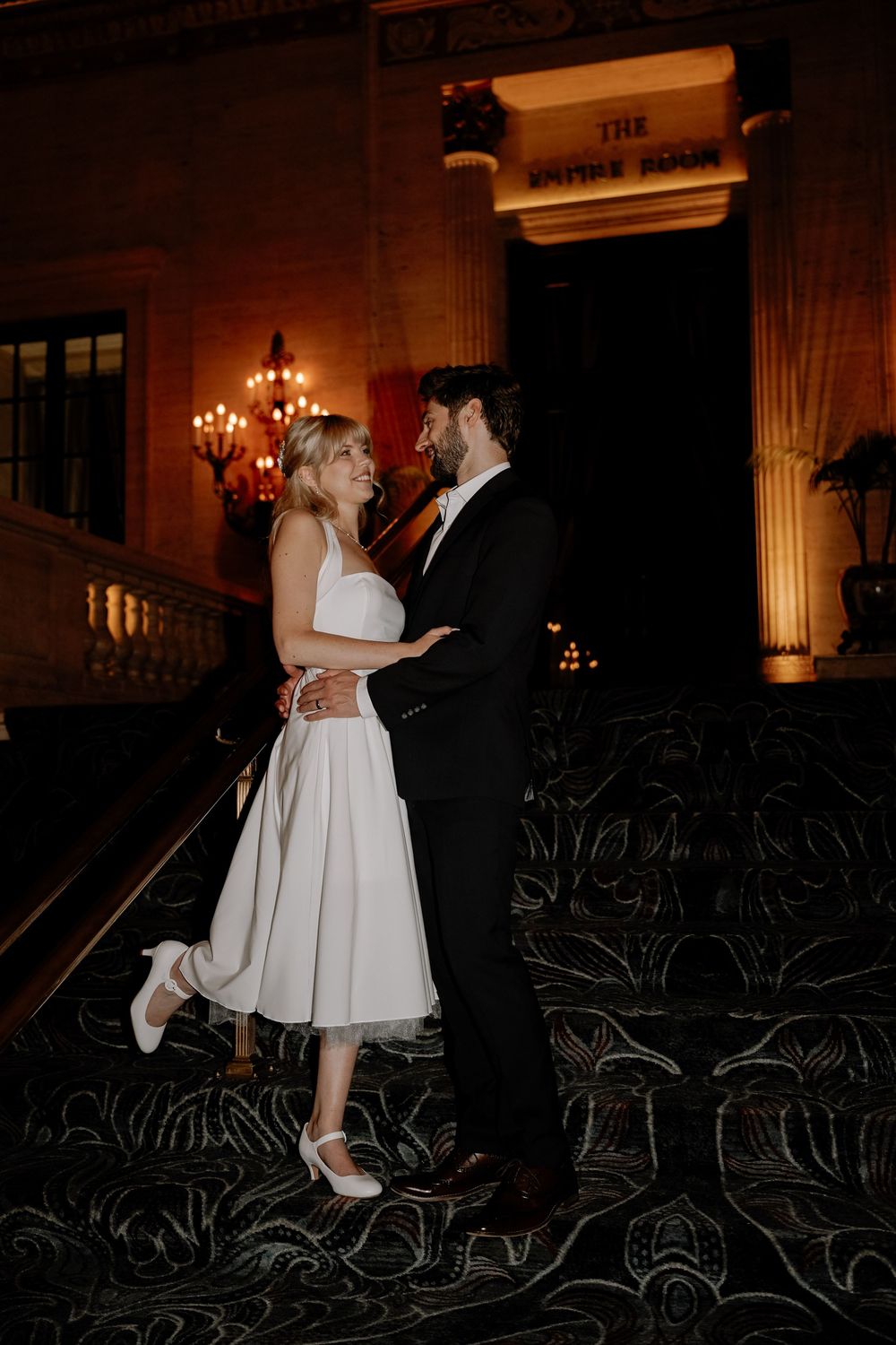 A couple shares an intimate moment on dark stairs with warm lighting and decorative railings in the background.
