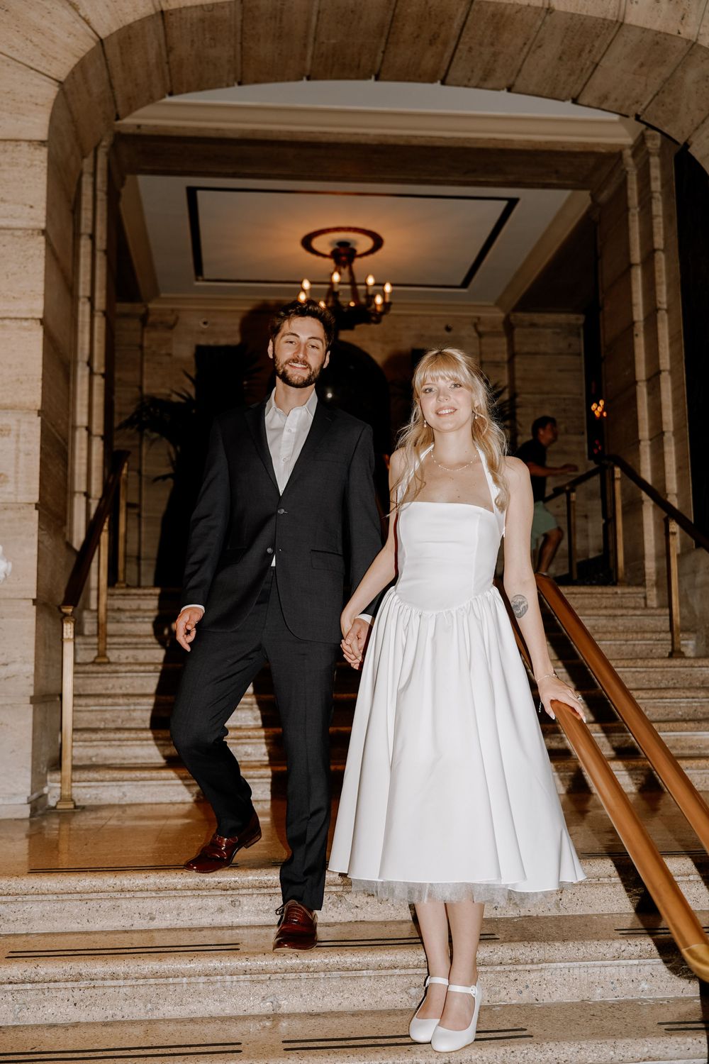 Couple dancing on grand marble staircase with chandelier visible in elegant indoor setting.