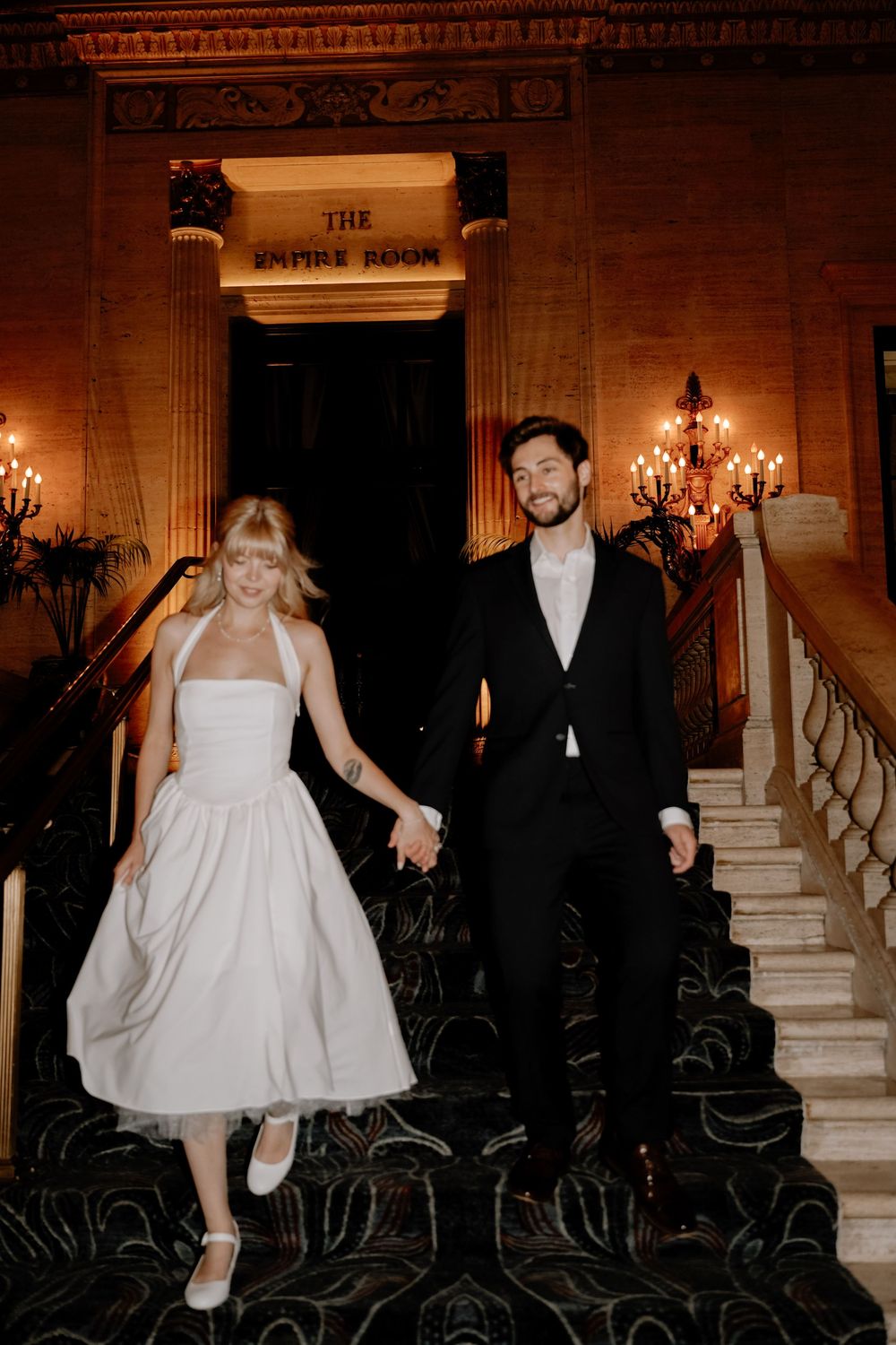 Romantic couple descends ornate staircase under vintage chandeliers in formal evening attire.