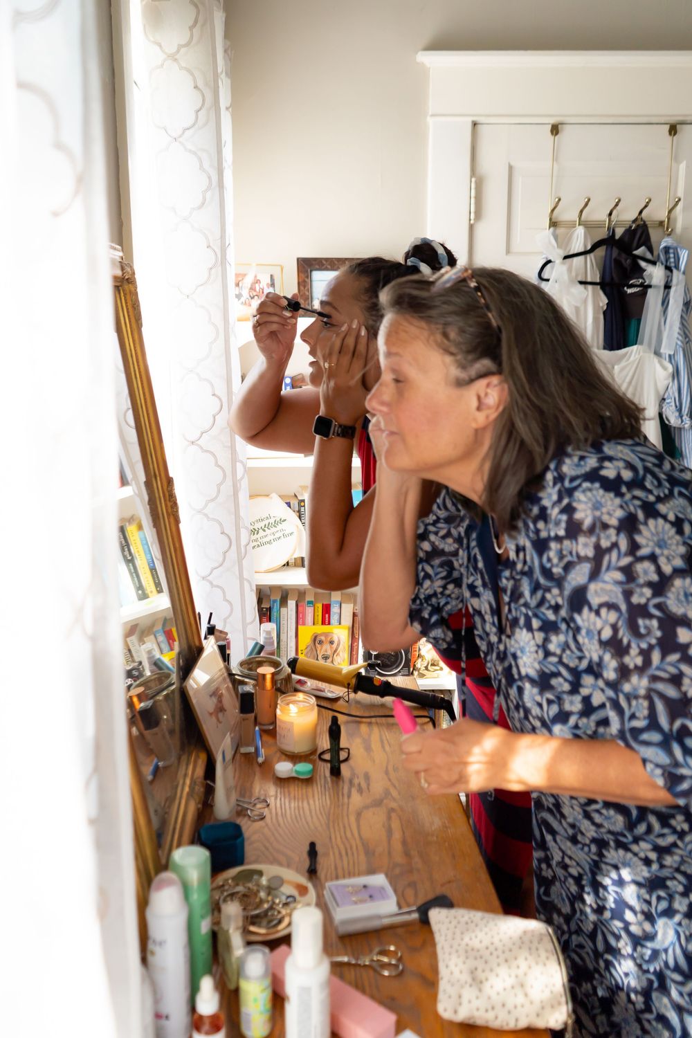 People applying makeup and getting ready at a wooden vanity with mirrors and beauty products scattered on the counter.