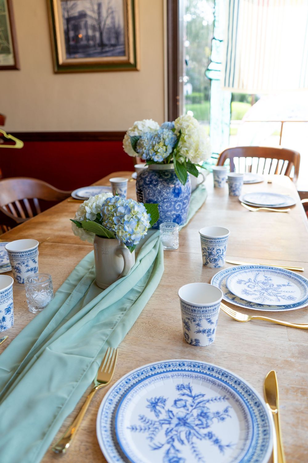Elegant table setting with blue and white china plates, sage green runners, and hydrangea centerpieces on wooden dining table.