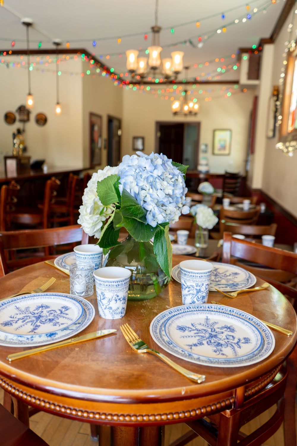 Close-up of elegant table setting with blue and white china plates, hydrangea centerpiece, and string lights in background for reception