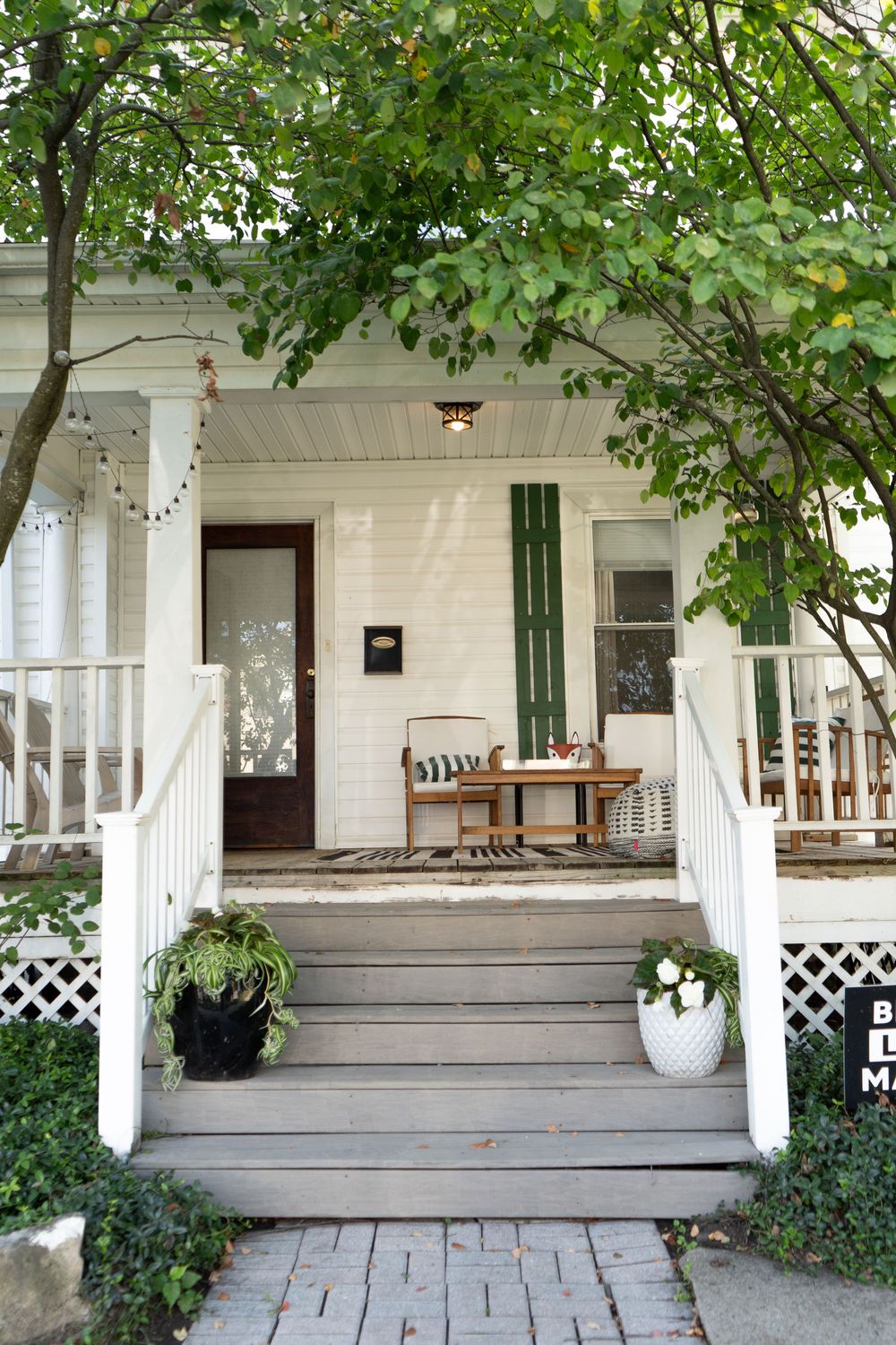 White porch with wooden steps, green lattice trim, and lush foliage creates a welcoming entrance to a classic home.